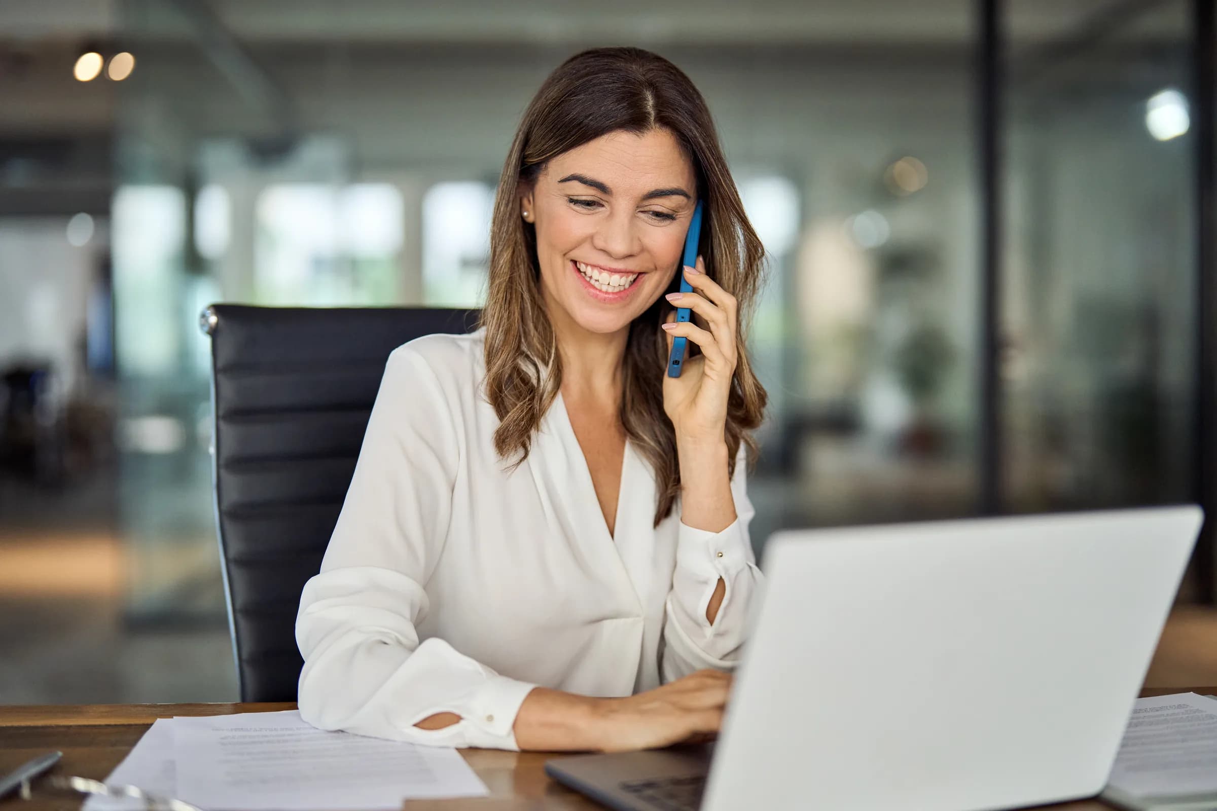 A recruitment consultant at her desk, smiling during a phone call, with a laptop open in a bright modern office